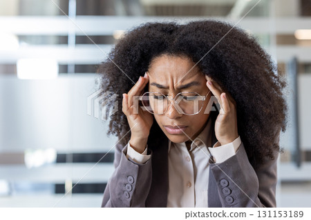 Close-up photo of a young African American woman in a business suit sitting at her workplace, eyes closed and holding her head in her hands. Close-up photo of a young African American woman in a business suit sitting at her workplace, eyes closed and holding her head in her hands. 131153189