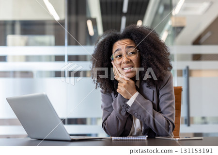 A sad African American woman sits at a desk in the office and holds her cheek with her hand, feeling a severe toothache. A sad African American woman sits at a desk in the office and holds her cheek with her hand, feeling a severe toothache. 131153191