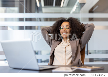 A satisfied and relaxed young African American woman in a suit is sitting in the office at a desk, hands behind her head and resting with her eyes closed. 131153357