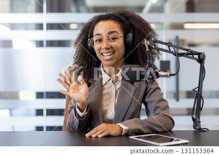 Smiling young African American woman sitting in office at desk wearing headphones in front of stack with microphone, talking and greeting to camera. Smiling young African American woman sitting in office at desk wearing headphones in front of stack with microphone, talking and greeting to camera. 131153364