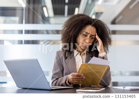 Upset young African American woman sitting at the desk in the office, holding her head with her hand and reading received letter with news and documents. 131153405