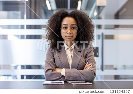 Portrait of a young serious African American business woman in a suit and glasses crossing her arms on her chest, sitting at the desk in the office and looking confidently at the camera. Portrait of a young serious African American business woman in a suit and glasses crossing her arms on her chest, sitting at the desk in the office and looking confidently at the camera. 131154397