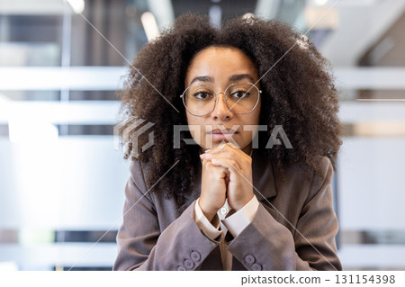 Close-up portrait of a young African-American woman in a suit and glasses sitting inside an office, holding her hands together and looking seriously at the camera. 131154398