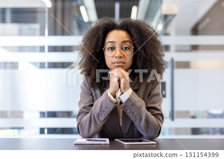 Close-up portrait of a young gray-haired African-American businesswoman in a suit and glasses sitting in the office at a desk, hands folded together and looking at the camera. Close-up portrait of a young gray-haired African-American businesswoman in a suit and glasses sitting in the office at a desk, hands folded together and looking at the camera. 131154399