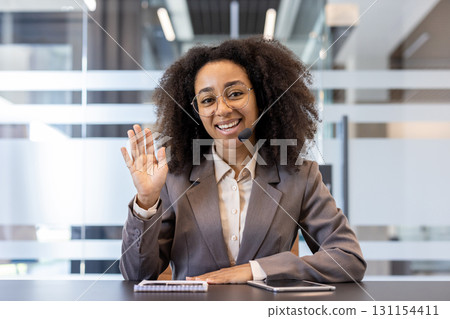 Smiling young African American woman wearing headset and suit sitting in office at desk, talking and greeting to camera. Smiling young African American woman wearing headset and suit sitting in office at desk, talking and greeting to camera. 131154411