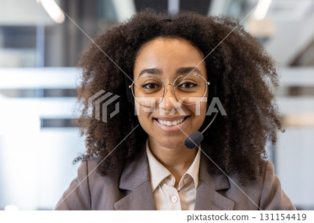 Portrait of a smiling young African American woman in a suit and glasses sitting in the office wearing a headset, looking confidently at the camera. 131154419
