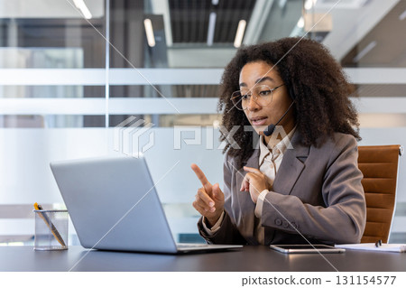 Young serious African-American woman in headset sitting in office at laptop and conducting online consultation, explaining and gesturing with hands to monitor. Young serious African-American woman in headset sitting in office at laptop and conducting online consultation, explaining and gesturing with hands to monitor. 131154577