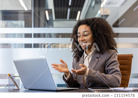 Smiling young African American woman in suit and headset sitting at desk in office and gesturing with hands talking on video call on laptop. Smiling young African American woman in suit and headset sitting at desk in office and gesturing with hands talking on video call on laptop. 131154578