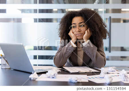 Tired and upset African American woman sitting in the office at the desk among crumpled paper and looking at the laptop screen. Tired and upset African American woman sitting in the office at the desk among crumpled paper and looking at the laptop screen. 131154594