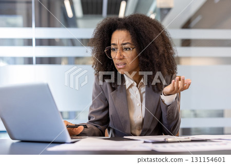 Frustrated young african american business woman sitting in office at work desk with documents and looking at laptop screen in frustration with arms outstretched. 131154601