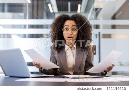 Portrait of disappointed and upset young African-American woman sitting in the office at the desk, holding documents and papers, throwing up her hands in frustration. Portrait of disappointed and upset young African-American woman sitting in the office at the desk, holding documents and papers, throwing up her hands in frustration. 131154610
