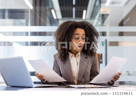 Confused young African American woman in a suit sitting in a business suit in the office and looking shocked at documents and reports in her hands. Confused young African American woman in a suit sitting in a business suit in the office and looking shocked at documents and reports in her hands. 131154611