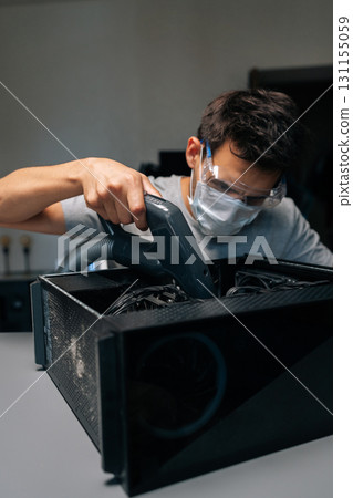 Vertical portrait of computer technician wearing protective mask and glasses using vacuum cleaner to clean inside of desktop computer case, performing PC maintenance and ensuring optimal performance. Vertical portrait of computer technician wearing protective mask and glasses using vacuum cleaner to clean inside of desktop computer case, performing PC maintenance and ensuring optimal performance. 131155059