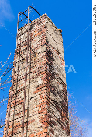 A weathered brick chimney rises against a blue sky, supported by a metal ladder 131155068