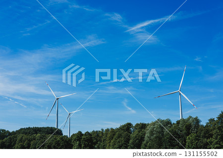 Wind turbines stand tall against a bright blue sky with wispy clouds in a serene landscape of green trees during a sunny day 131155502