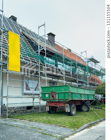 Bright yellow banner highlights house renovation project with scaffolding and a green trailer parked nearby in a quiet neighborhood Bright yellow banner highlights house renovation project with scaffolding and a green trailer parked nearby in a quiet neighborhood 131155504