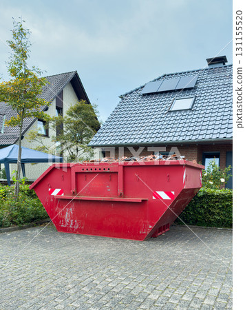 Large red dumpster filled with debris placed outside a modern home on a cloudy day in a suburban neighborhood 131155520