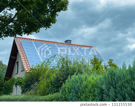 Modern home adorned with solar panels under a cloudy sky surrounded by lush greenery in a rural setting during early afternoon 131155530