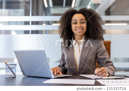 Portrait of a smiling young African-American businesswoman working in the office, sitting at a desk with a laptop, making notes in a notebook and looking at the camera. 131155718