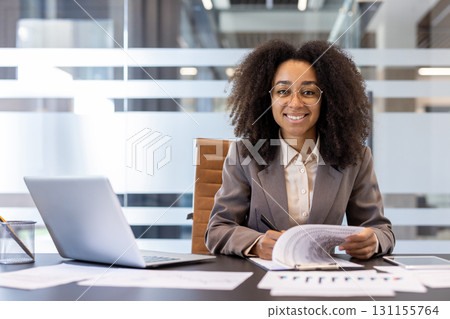 Portrait of a young smiling African-American businesswoman sitting in the office at the table with a laptop and documents, signing an agreement and contract, looking confidently at the camera. Portrait of a young smiling African-American businesswoman sitting in the office at the table with a laptop and documents, signing an agreement and contract, looking confidently at the camera. 131155764