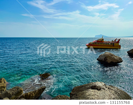 Colorful boat sails through clear blue water near rocky coastline with distant mountains and sunny sky in the background Colorful boat sails through clear blue water near rocky coastline with distant mountains and sunny sky in the background 131155768