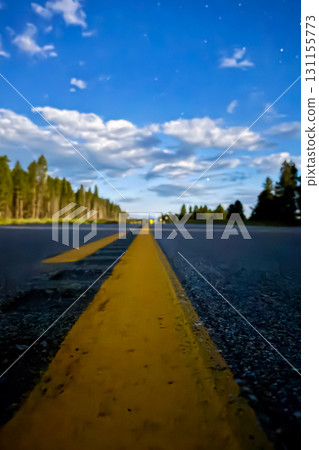 Stretching yellow lines lead down a wide open road under a bright blue sky filled with fluffy clouds during golden hour before dusk 131155773