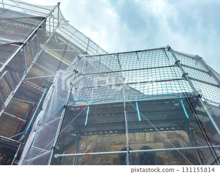 Repair work in progress on historic building under cloudy sky, showcasing scaffolding and protective netting Repair work in progress on historic building under cloudy sky, showcasing scaffolding and protective netting 131155814