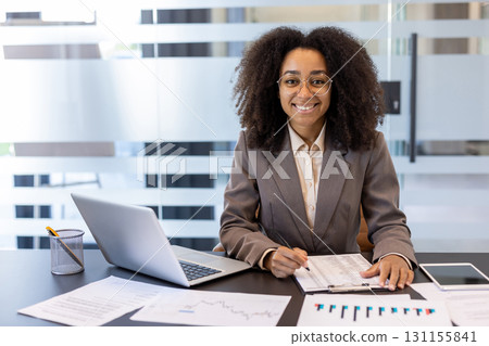 Portrait of a young curly-haired African-American woman in a suit and glasses working in the office with a notebook and documents, looking at the camera with a smile. 131155841