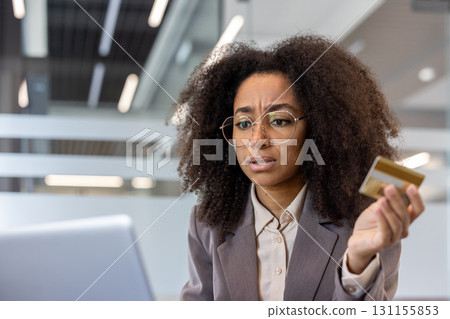 Upset young African American woman sitting at desk in office, holding credit card and looking frustrated at laptop screen. Upset young African American woman sitting at desk in office, holding credit card and looking frustrated at laptop screen. 131155853