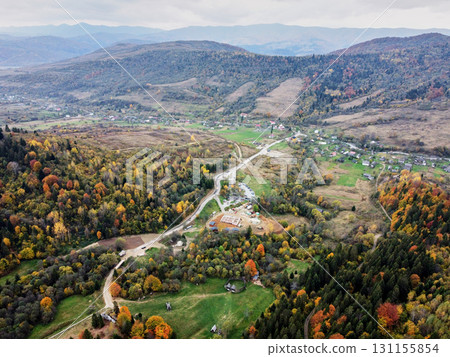 Aerial view of tranquil valley in autumn. Vibrant fall foliage and small village nestled among rolling hills. Winding roads connect scattered houses. Carpathian mountains, Ukraine. Aerial view of tranquil valley in autumn. Vibrant fall foliage and small village nestled among rolling hills. Winding roads connect scattered houses. Carpathian mountains, Ukraine. 131155854