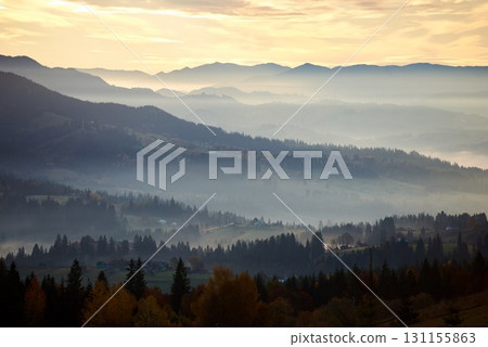 Misty autumn morning in mountain valley. Layers of hills bathed in soft light, with patches of sunlight breaking through fog, illuminating autumn foliage. Interplay of light and shadow. 131155863