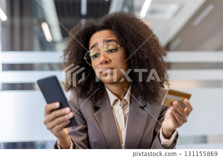 Close-up photo of a sad young African-American woman sitting at a desk in the office, holding a credit card and looking frustrated at the phone screen. Close-up photo of a sad young African-American woman sitting at a desk in the office, holding a credit card and looking frustrated at the phone screen. 131155869