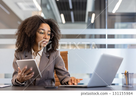 Young African American businesswoman multitasking with laptop and tablet. Professional woman focused on her work in modern office environment. Concept of productivity, technology, and business. Young African American businesswoman multitasking with laptop and tablet. Professional woman focused on her work in modern office environment. Concept of productivity, technology, and business. 131155984