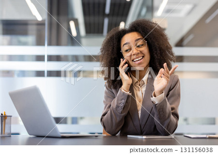 Smiling and successful young African American business woman sitting in the office at the desk and talking on the phone while gesturing with her hands. 131156038