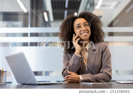 Smiling young African-American businesswoman sitting in office in suit at desk and talking on mobile phone. Smiling young African-American businesswoman sitting in office in suit at desk and talking on mobile phone. 131156042