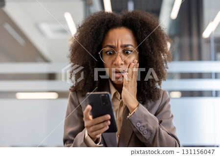 Young African American businesswoman looking shocked at smartphone in modern office environment. Woman appears surprised by unexpected news or message on phone while working 131156047