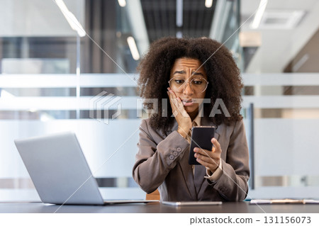 Shocked and scared young African American woman is in the office, sitting at the desk and looking at the mobile phone screen, holding her hand to her cheek. 131156073