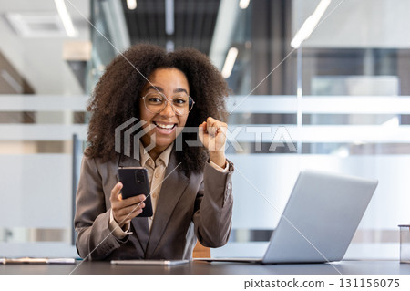 Portrait of young African American businesswoman who received good news and result, sitting in office at desk, holding phone, raised victory fist up and looking smiling at camera. Portrait of young African American businesswoman who received good news and result, sitting in office at desk, holding phone, raised victory fist up and looking smiling at camera. 131156075