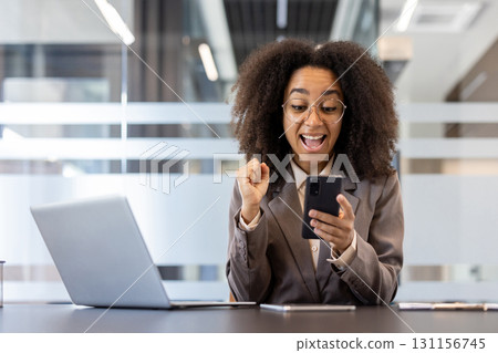 Young African American businesswoman in formal attire celebrating success at workplace inside office holding phone with laptop in front showing excitement and joy Young African American businesswoman in formal attire celebrating success at workplace inside office holding phone with laptop in front showing excitement and joy 131156745