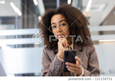 Young African American businesswoman in business attire using phone in office. Confident professional executive working and thinking about business tasks. Modern workplace environment. Young African American businesswoman in business attire using phone in office. Confident professional executive working and thinking about business tasks. Modern workplace environment. 131156752