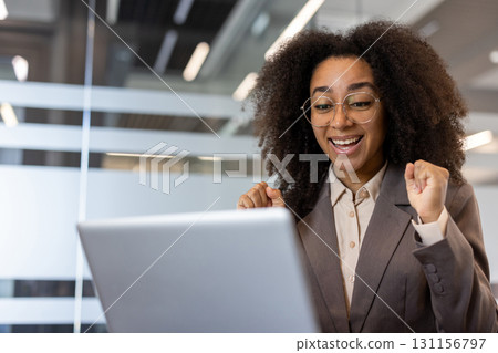 Happy young African American woman in business suit sitting in office at desk in front of laptop and enjoying achievement. Happy young African American woman in business suit sitting in office at desk in front of laptop and enjoying achievement. 131156797