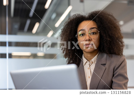 Close-up portrait of a young African-American woman in a business suit sitting in an office at a desk in front of a laptop, looking seriously at the camera. 131156828