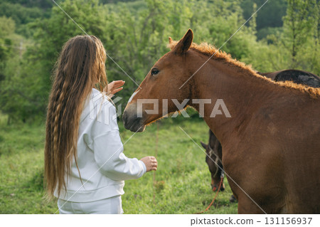 A woman is petting a brown horse in a field. The horse is standing next to another horse. 131156937
