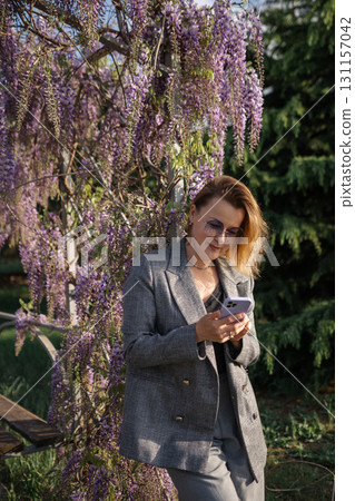 Woman, Phone, Garden - Woman in grey suit using her phone standing next to a blooming wisteria tree in a garden. Woman, Phone, Garden - Woman in grey suit using her phone standing next to a blooming wisteria tree in a garden. 131157042