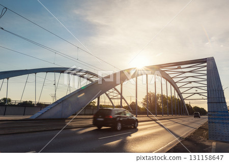 Scenic dynamic view of Magdeburg steel arch Jerusalem bridge cars crossing under warm sunrise sunlight. Urban composition highlights architectural structure traffic movement modern city atmosphere Scenic dynamic view of Magdeburg steel arch Jerusalem bridge cars crossing under warm sunrise sunlight. Urban composition highlights architectural structure traffic movement modern city atmosphere 131158647