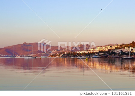 A stunning sunrise over a calm, reflective bay, with sailboats moored in the glassy water. The sun's first rays illuminate the mountains and the whitewashed buildings of a coastal town on the hillside 131161551