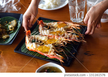 A close-up photo of a hand supporting a plate of large grilled shrimp that had been cut in half, showing oozing fat. Placed on a green banana leaf 131161576