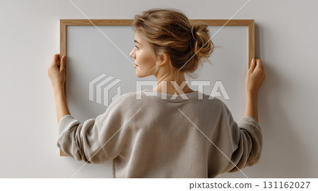 Young woman in a modern apartment hanging a large poster frame. Rear view of a girl with an empty blank frame on the wall, setting up a mockup for an artwork. 131162027