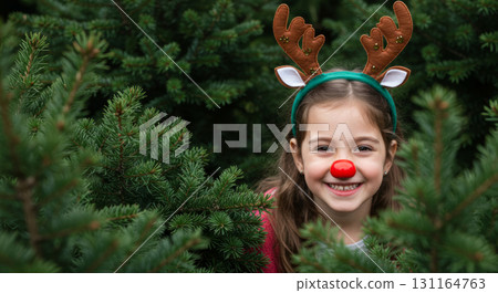 A cheerful girl in reindeer antlers and a red nose peeks from behind a Christmas tree, full of holiday spirit. 131164763