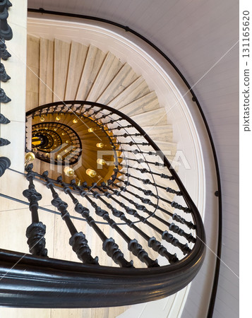 Spiral marble staircase with ornate black balustrade and warm hanging lights viewed from above 131165620